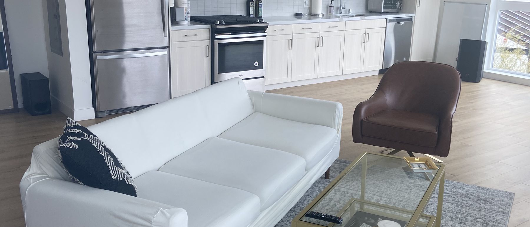 Los Angeles short-term rental living room after an Airbnb turnover: white sofa with cushions squared, glass coffee table cleared, rug straightened, open kitchen behind it wiped down and clear of dishes.